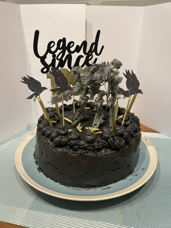 A picture of a cake with raven figurines and an Armored Core action figure on top, covered in jet black frosting.