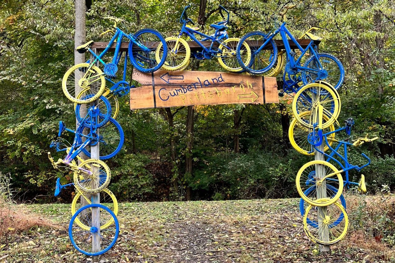 A wooden sign with blue and yellow bicycles attached. The sign text reads "Cumberland" and "Pittsburgh," with arrows pointing in opposite directions.