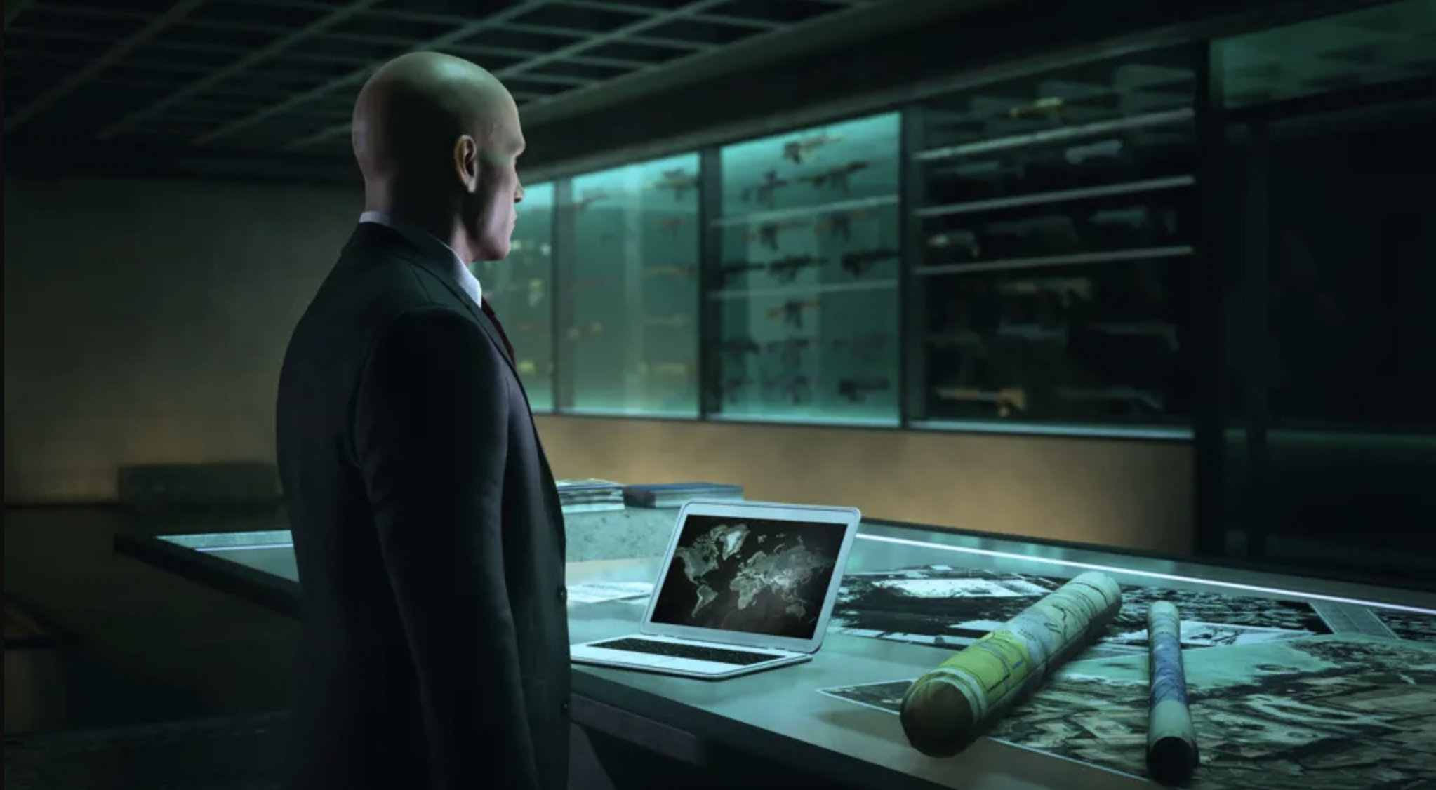 Agent 47, a bald white man in a suit, stands in front of a desk with a computer and rolled-up back. On the wall in front of him is a case full of weapons.