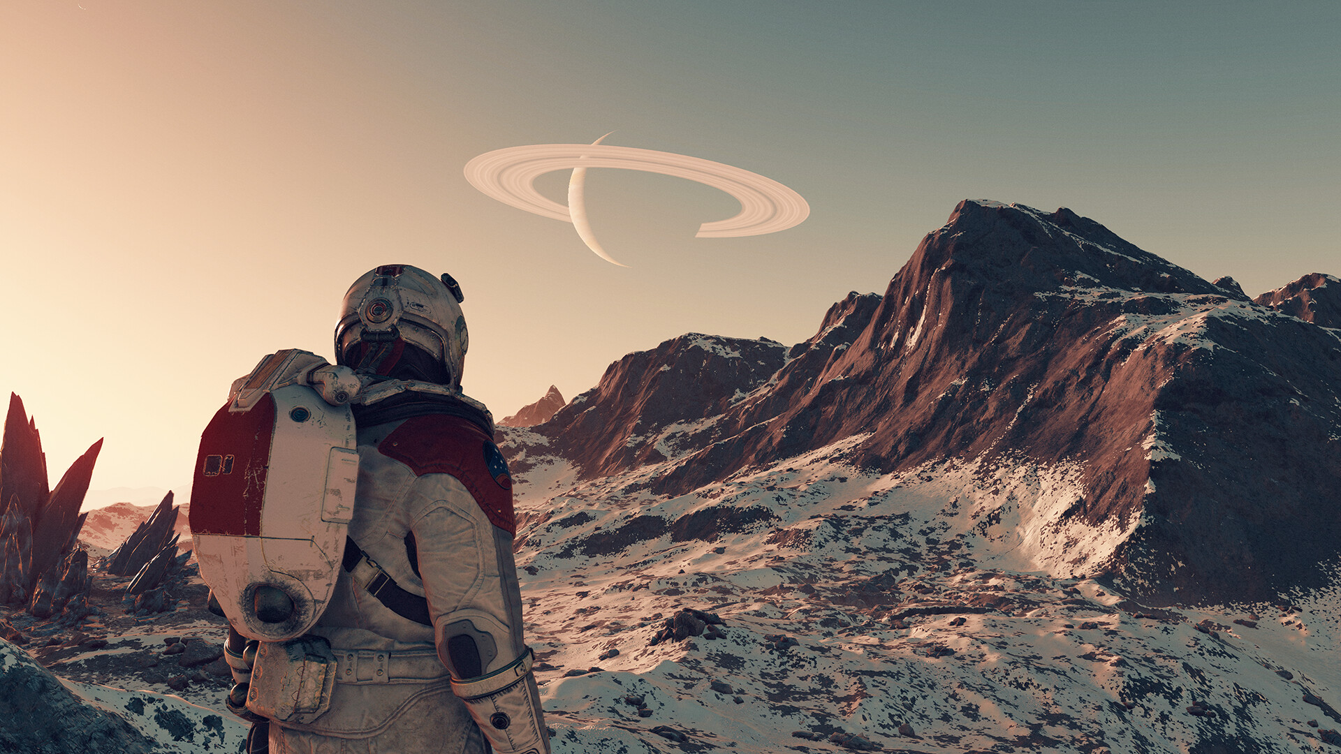 A screenshot from the video game Starfield: a figure in a space suit stands on a planet with snowy ground and mountains. In the sky there's the outline of a planet with a ring around it.
