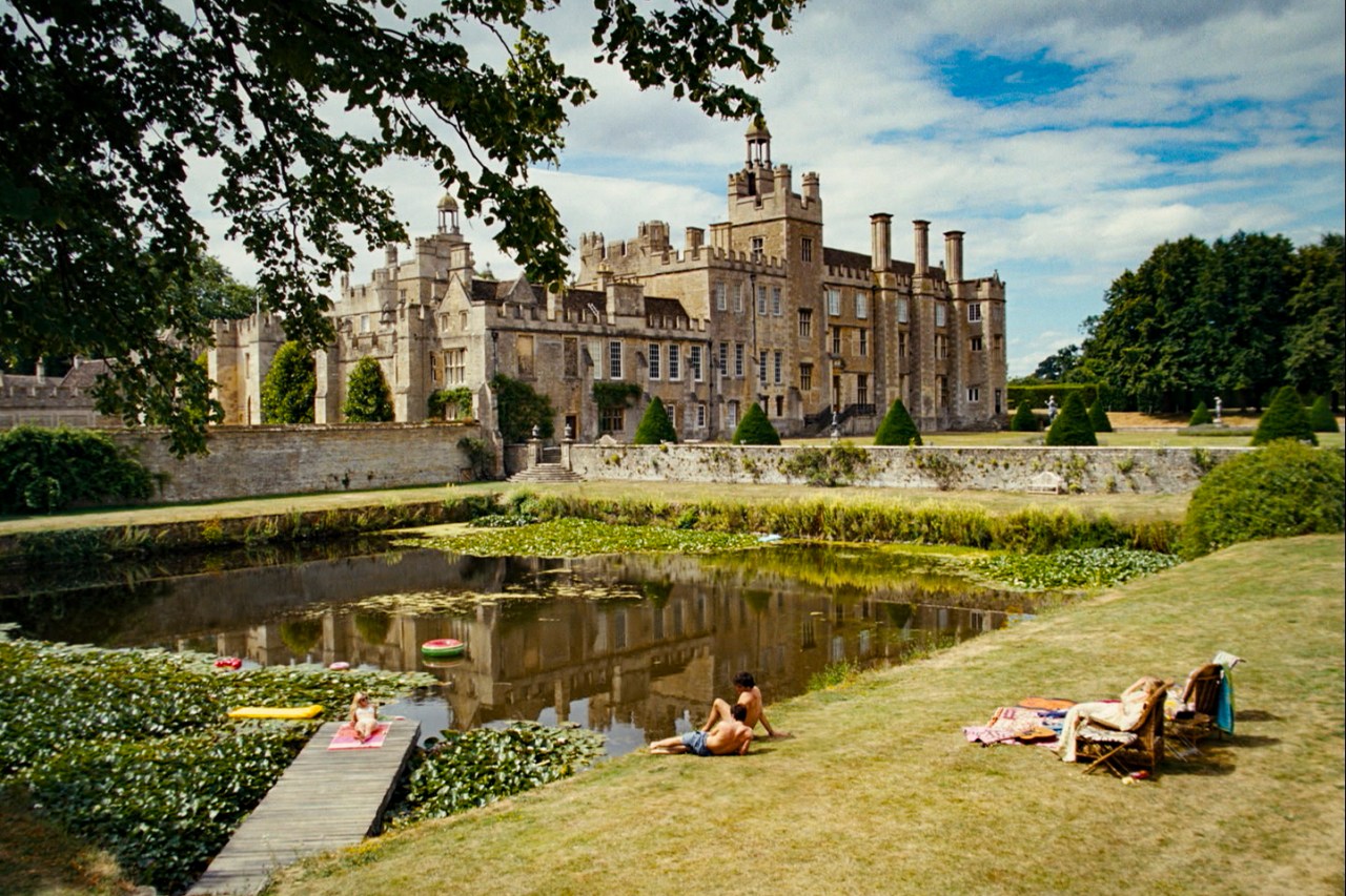 A screenshot from Saltburn. In a wide angle shot, you see the entirety of the Salturn estate and the part of the grounds that overlook an artificial lake. Three figures rest by the lakeside.