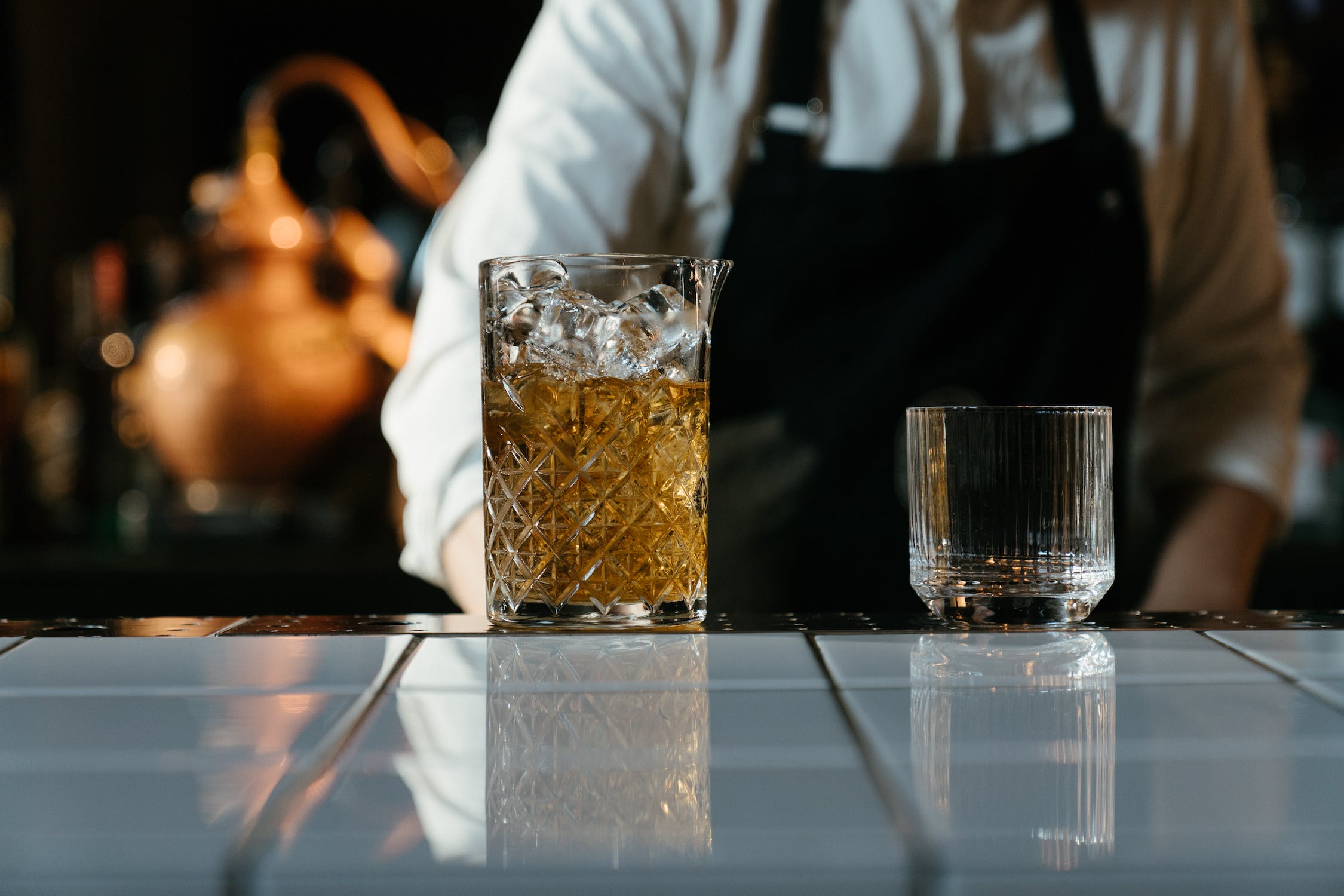 A clear glass with brown liquid on a white tile counter, next to an empty glass. A person in a white shirt and black apron stands behind them.