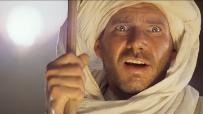 A screenshot from the movie "Indiana Jones and the Raiders of the Lost Ark:" Harrison Ford with a white scarf on his head, holding a stick