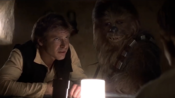 A screenshot from the movie "Star Wars: A New Hope:" Harrison Ford as Han Solo, wearing a white shirt and black vest, leaning over a table next to Chewbacca
