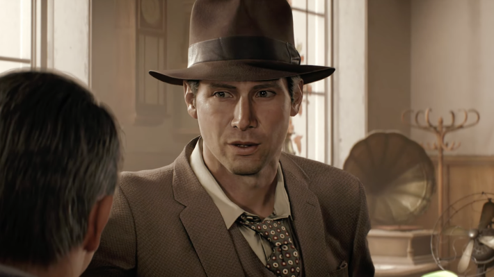 A screenshot from video game Indiana Jones and the Great Circle: Indiana Jones in a brown hat, brown suit coat, white shirt, and brown tie with white spots, standing in an office