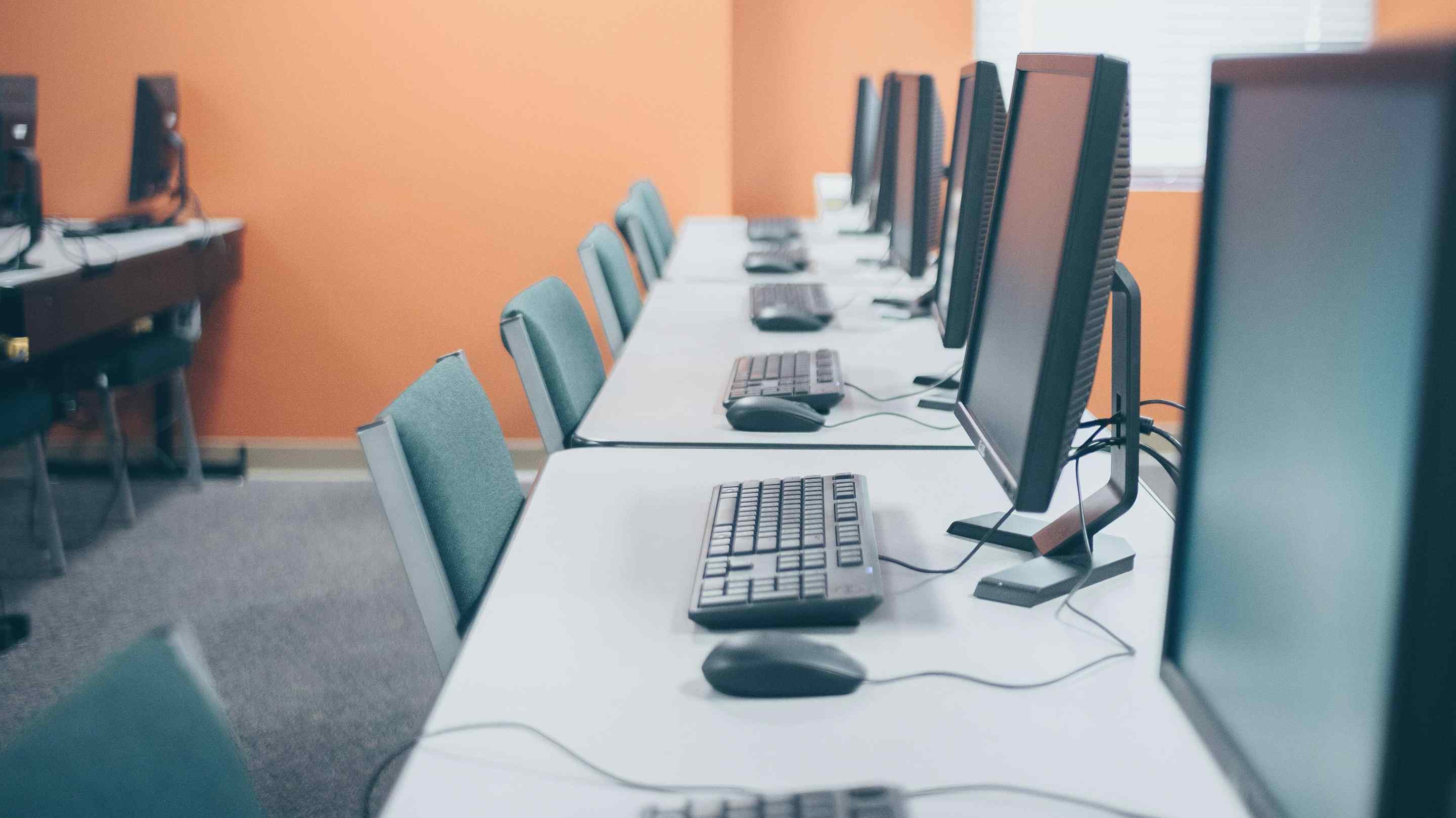 A row of computer monitors and keyboards on a white table, in front of empty chairs