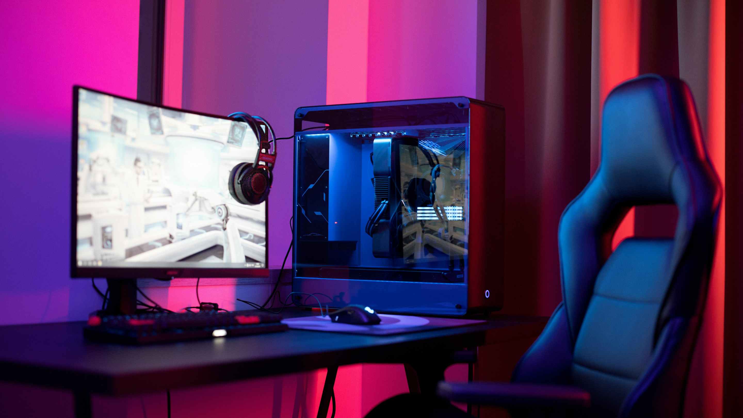 A gaming PC with a clear side sits on a desk, besides a glowing monitor and padded gaming chair