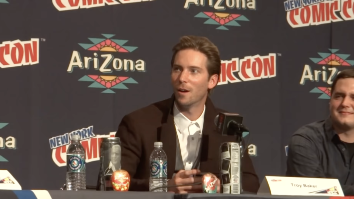An image of actor Troy Baker at New York Comic-Con: Baker, wearing a white shirt and black suit coat, sitting behind a table of microphones and water bottles