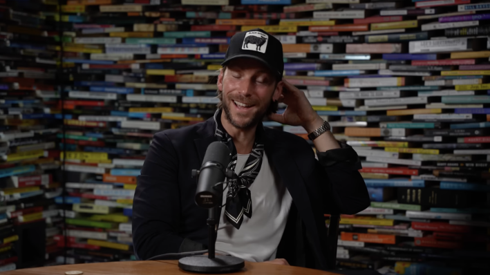 Actor Troy Baker on a podcast: wearing a white t-shirt, black shirt, and black baseball hat, in front of a wall of books