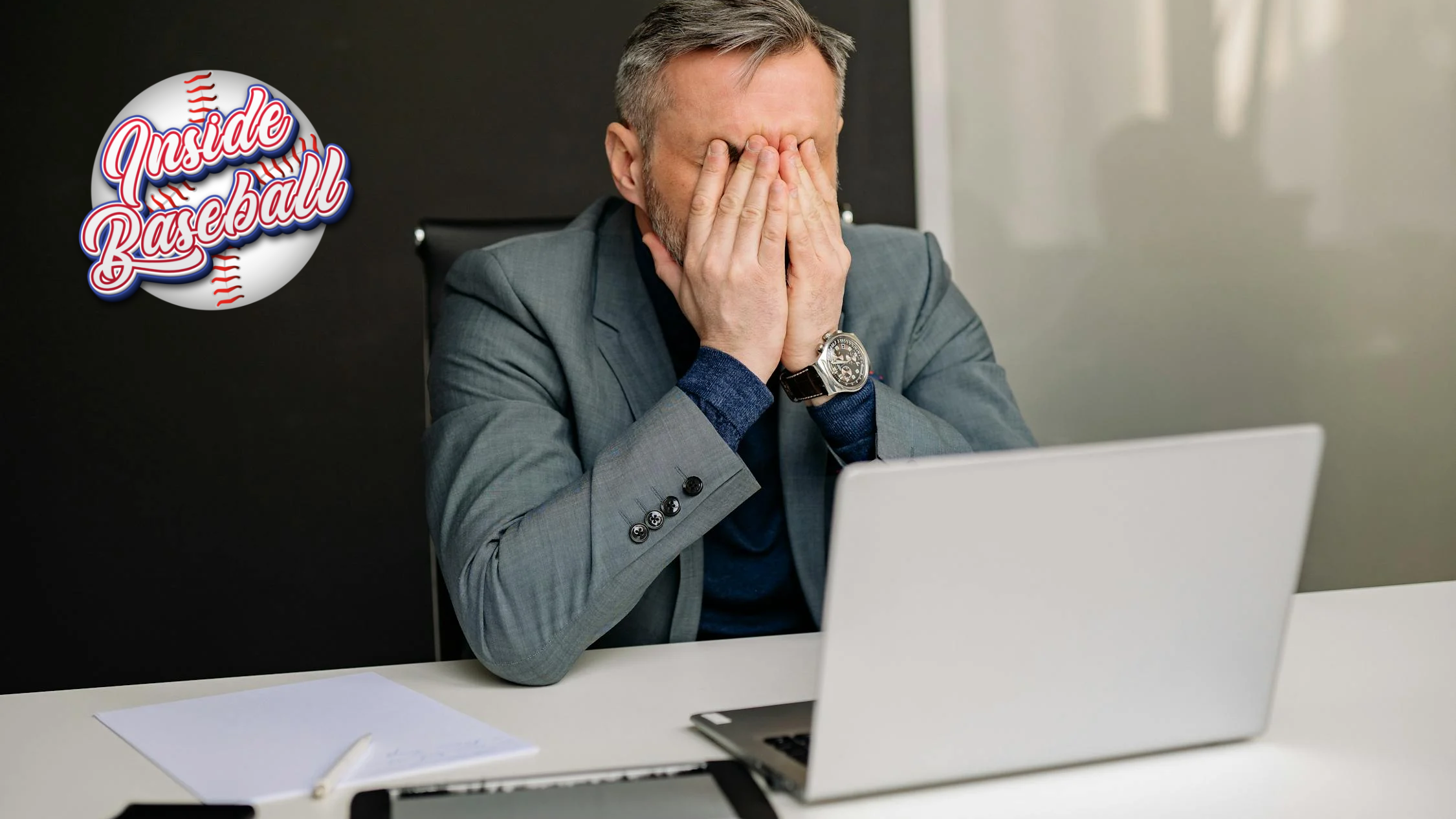 A man in a suit jacket sits in front of a laptop, holding his face in his hands