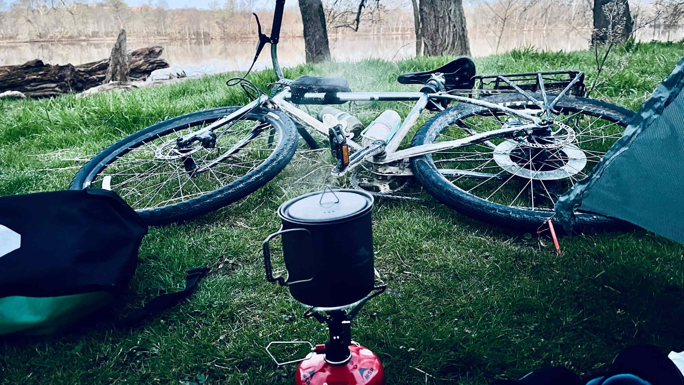 A photo of a bike on green grass in front of a river. In front of the bike is a camping stove with a small pot on top of it