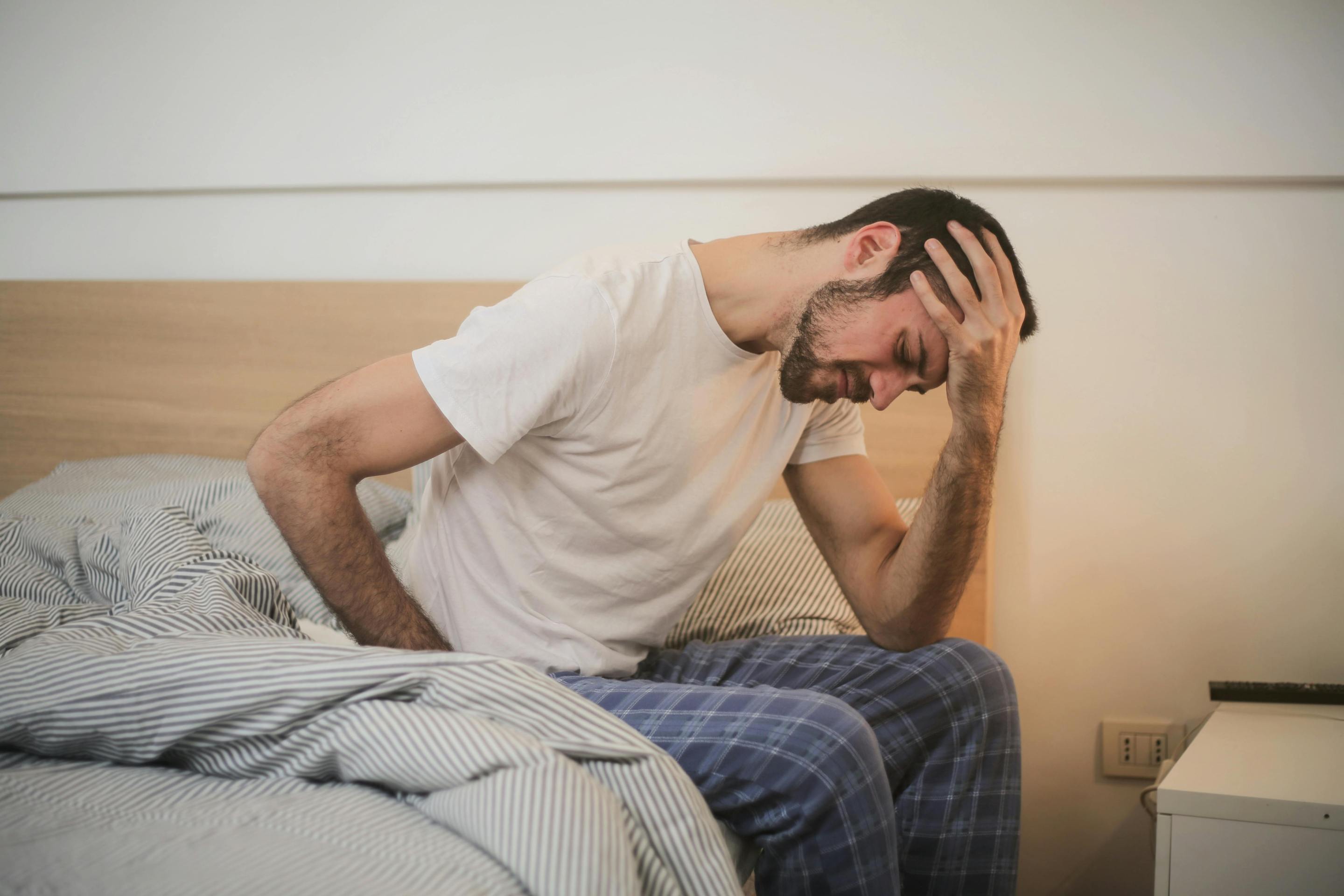 A man in a white t-shirt and blue pants sits on a bed, holding one hand to his forehead