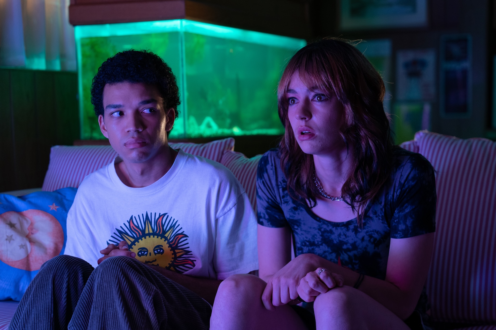 A screenshot from "I Saw The TV Glow:" two tween characters sit on a couch. One stares engrossed at something off-screen; the other stares at her.