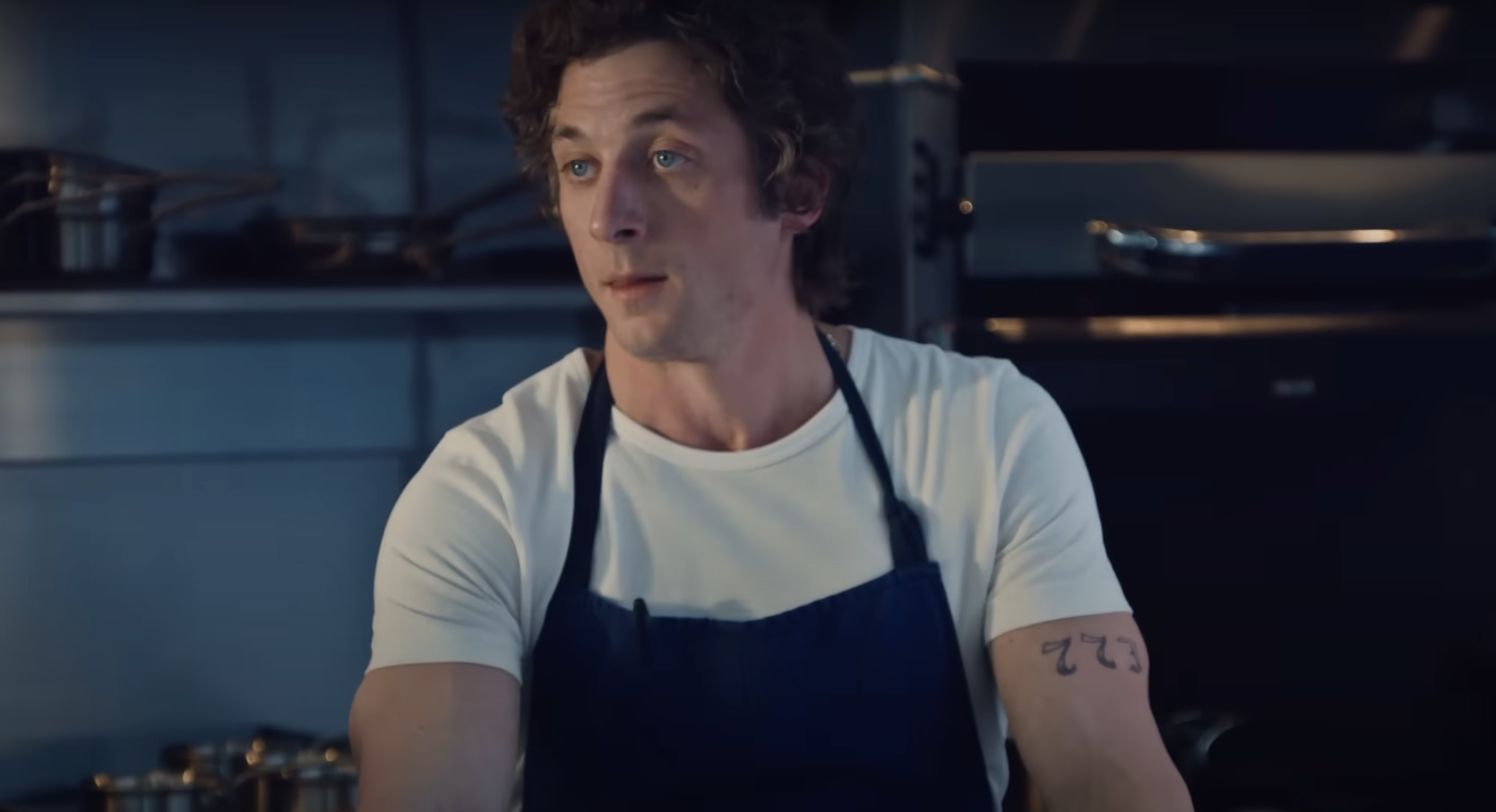 A screenshot from "The Bear:" main character Carmy, in a white tshirt and blue apron, stands behind a counter