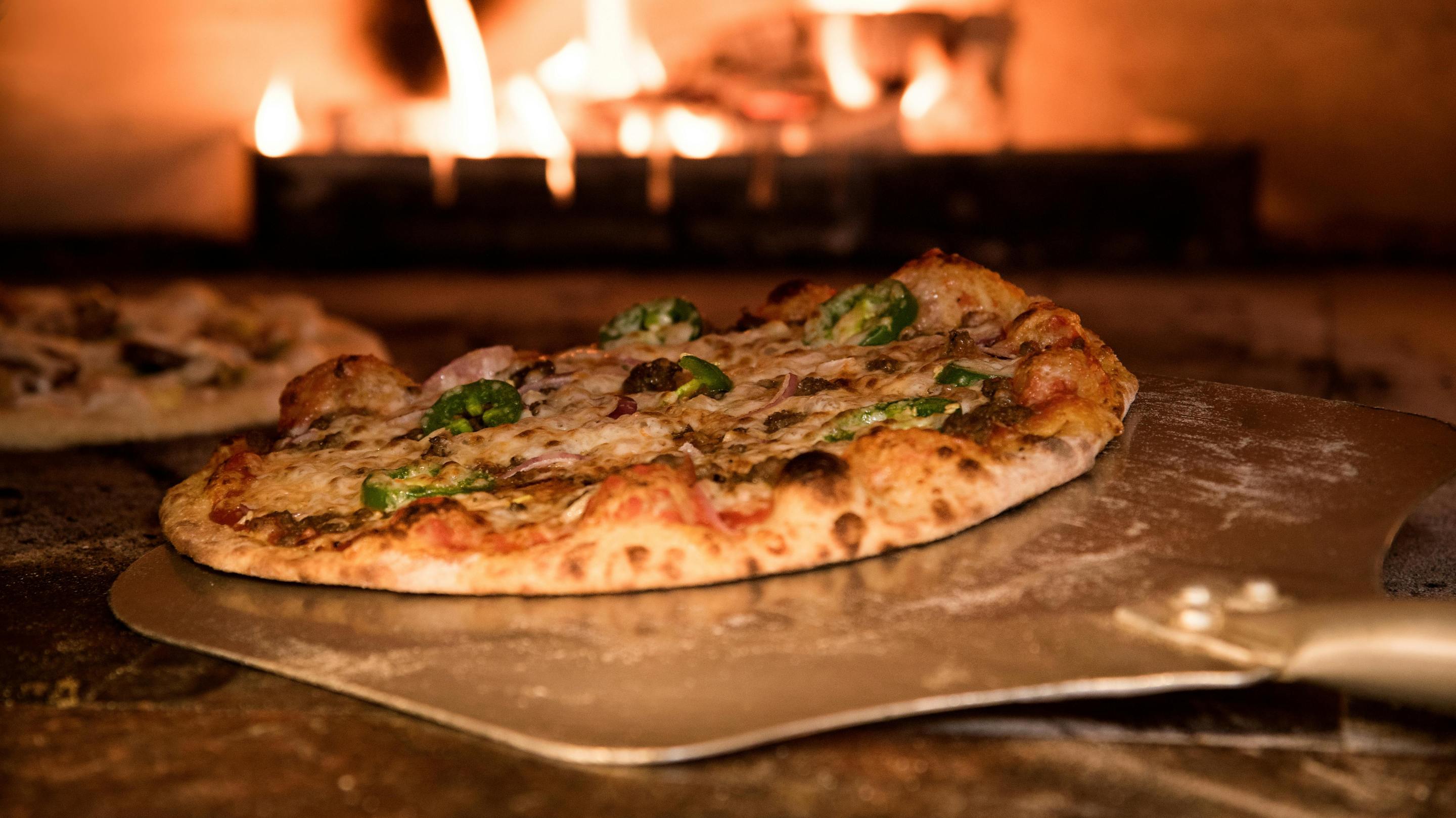 A pizza on a metal peel, in front of a burning oven