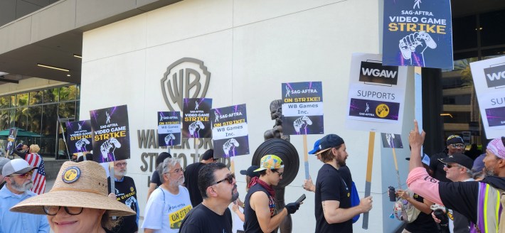 Video game performers picket outside of WB Studios in Burbank, holding signs that read "SAG-AFTRA on strike"