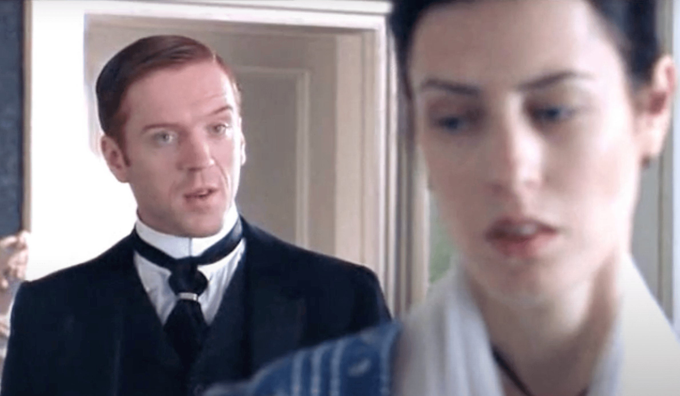 A screenshot from The Forsyte Saga: a man with red hair stares at a dark-haired woman from the background