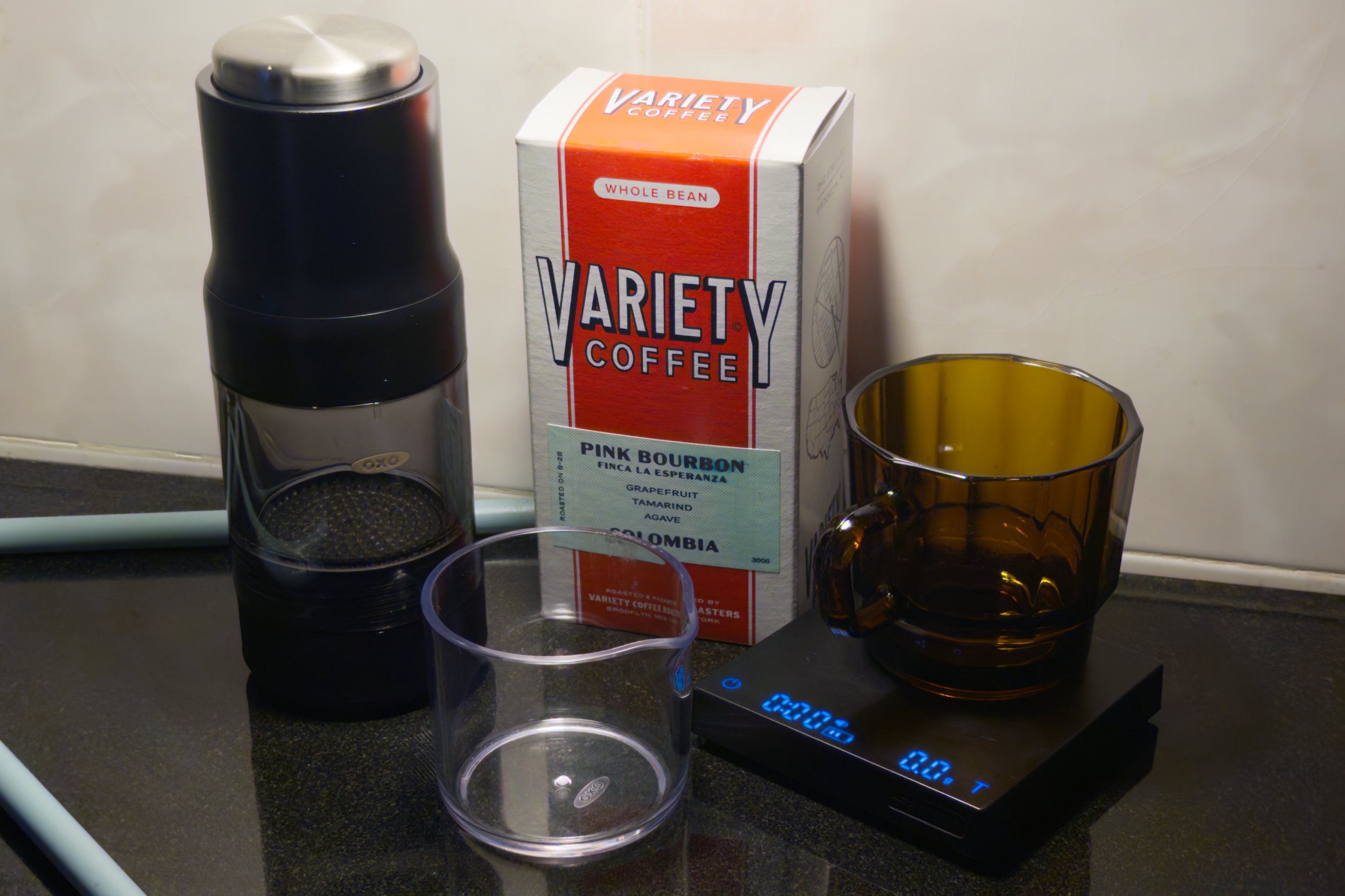 The OXO Brew Rapid Brewer on a table, next to a Variety Coffee box labeled "pink bourbon" and a scale is nearby tare'd to O. There is a glass on the scale made in Taiwan from recycled cellphone screens apparently. An empty Carafe that's included is in the foreground.