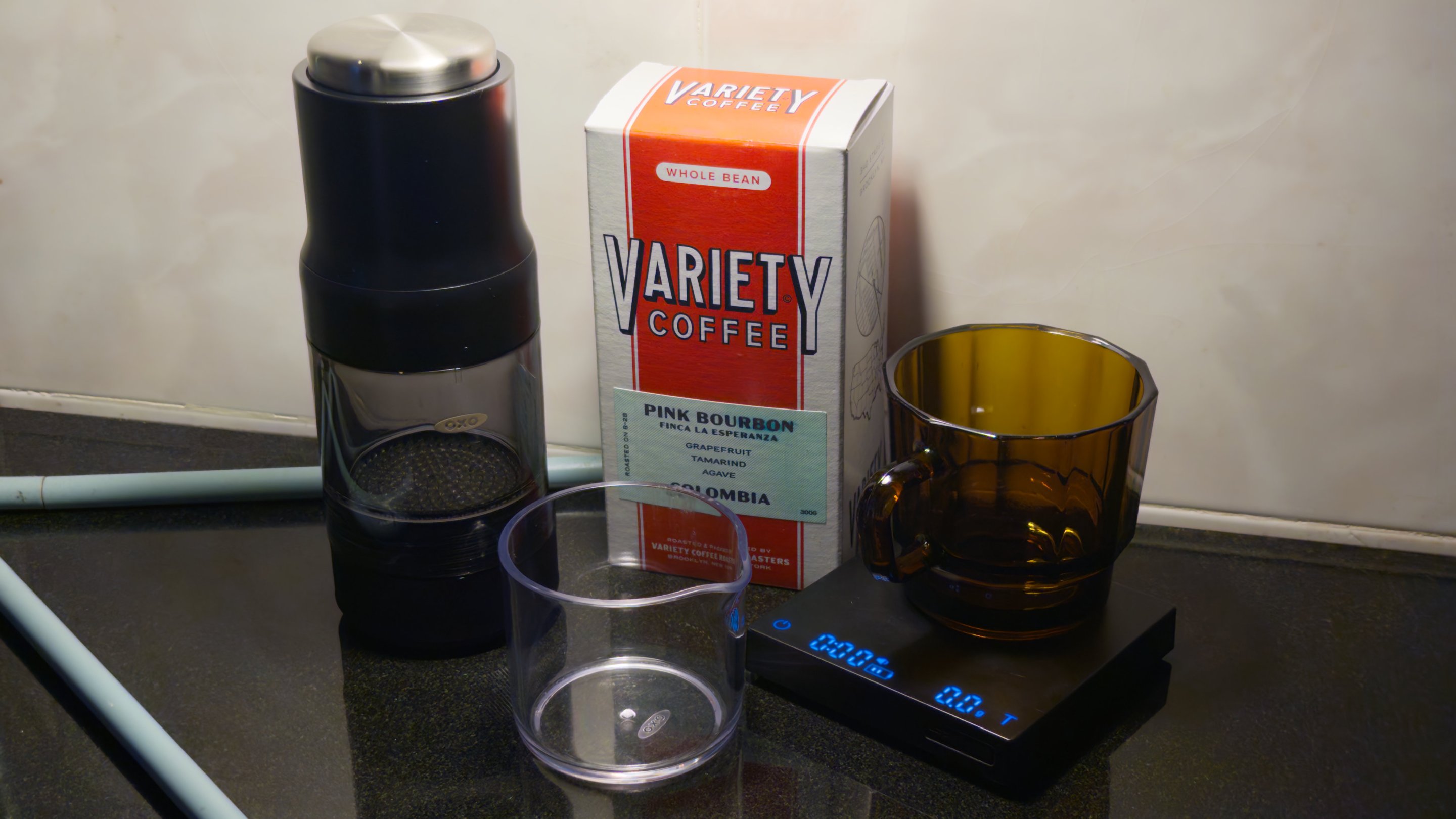 The OXO Brew Rapid Brewer on a table, next to a Variety Coffee box labeled "pink bourbon" and a scale is nearby tare'd to O. There is a glass on the scale made in Taiwan from recycled cellphone screens apparently. An empty Carafe that's included is in the foreground.